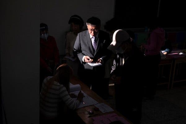 A man in a suit holding a piece of paper and standing in a dim room in which a ray of light falls on him.